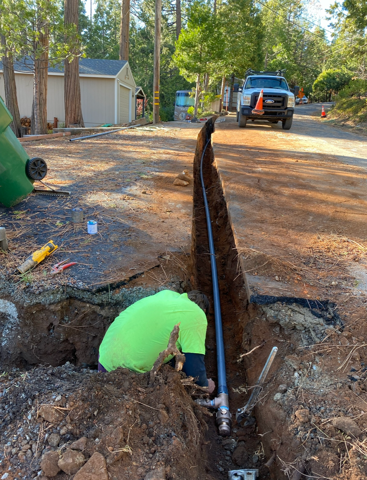A worker in a neon green shirt kneels in a trench to install a pipe in a wooded, residential area with a truck parked nearby.