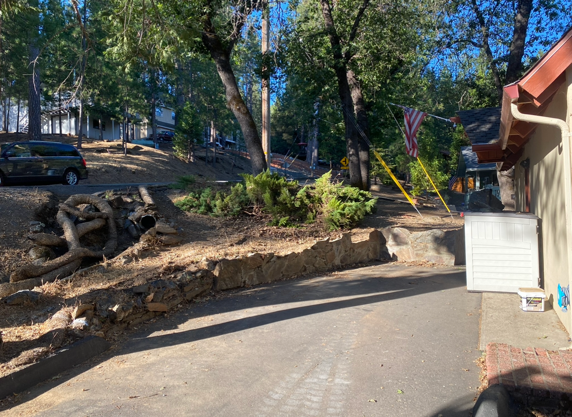 A paved driveway leads toward a house with a stone retaining wall, large trees, and an American flag in the background.