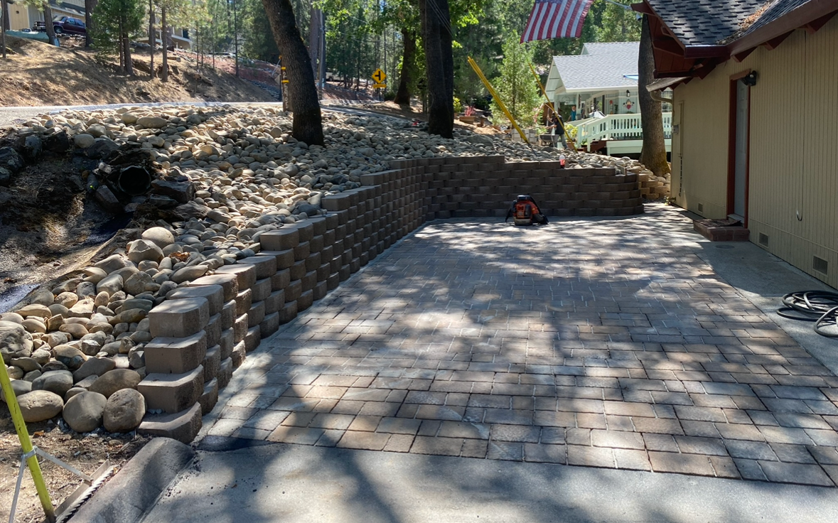 Brick patio with retaining walls. Gravel slope and trees in the background. Sunny day.