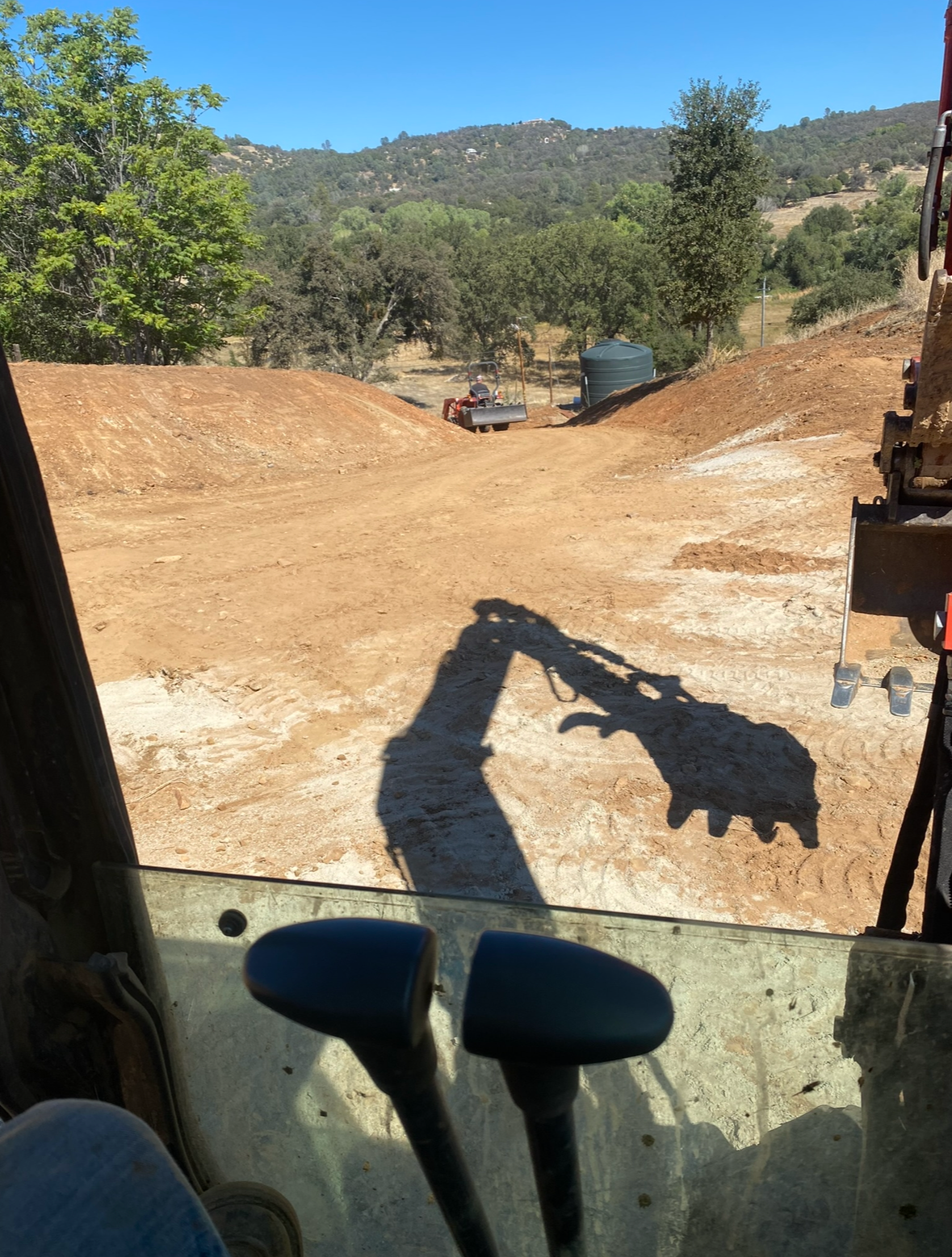 View from inside an excavator cab overlooking a dirt excavation site with a hill, trees, and a distant tractor.