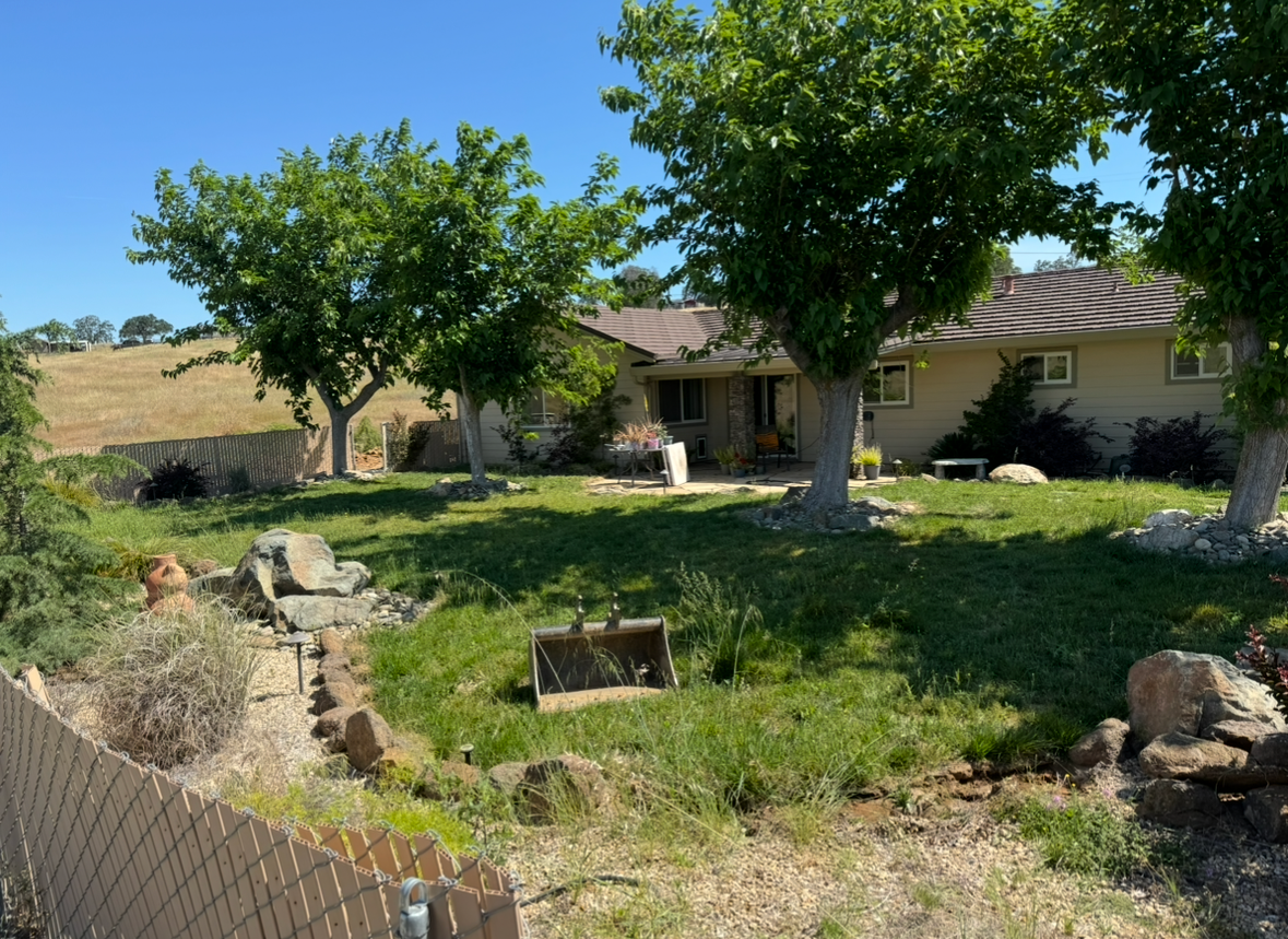A tan one-story house with a brown roof, surrounded by green trees, a grassy yard, and rocky landscaping on a sunny day.