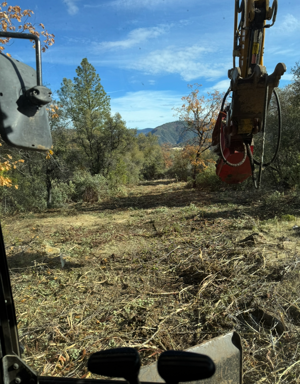 A view from inside heavy machinery cab, overlooking a cleared, brush-covered field with hills under a blue sky.