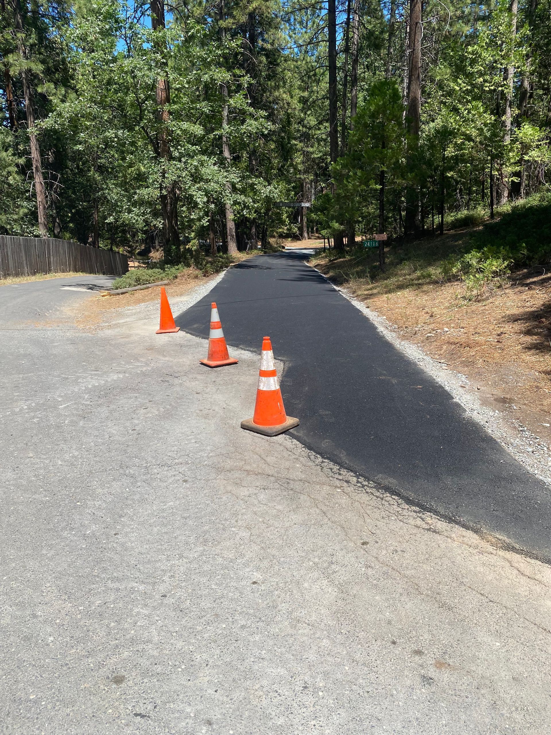 Three orange traffic cones line the edge of a newly paved black asphalt path leading into a wooded area.