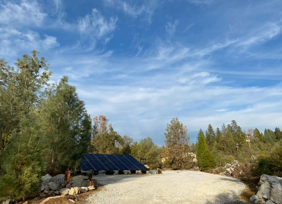 Solar panels installed on a metal frame in a graveled outdoor area surrounded by trees under a bright blue sky.