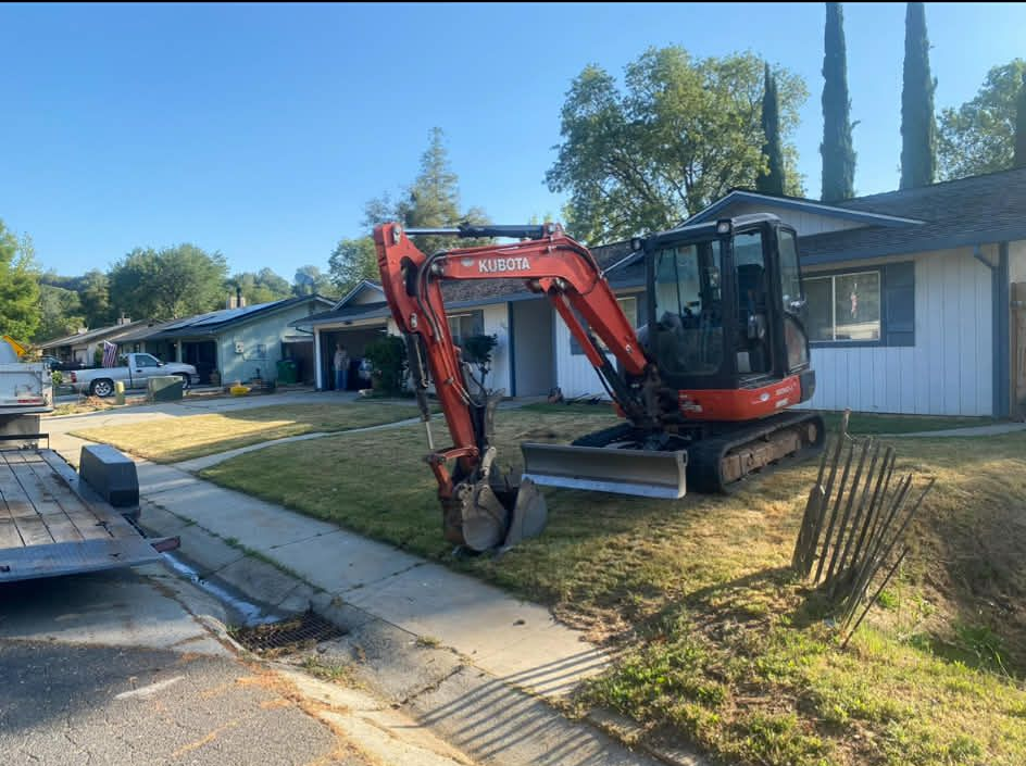 An orange Kubota excavator sits on the front lawn of a suburban house on a sunny day next to a trailer.