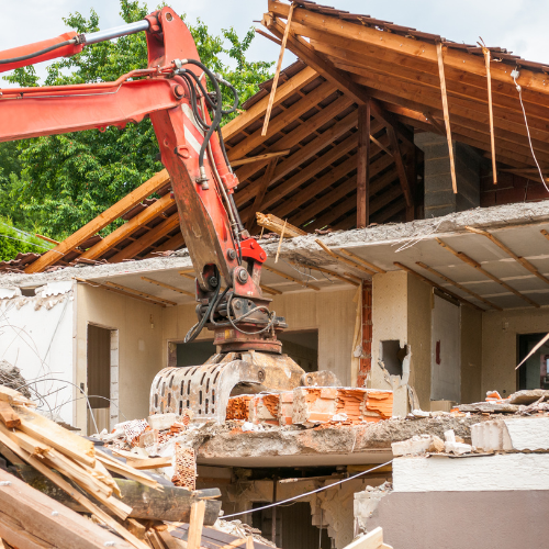 A red excavator is demolishing a house with a wooden roof.