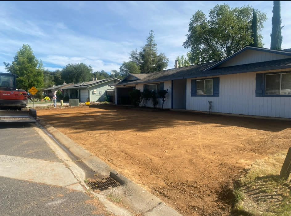 A construction vehicle parked next to a house with a newly cleared dirt front yard under a clear blue sky.