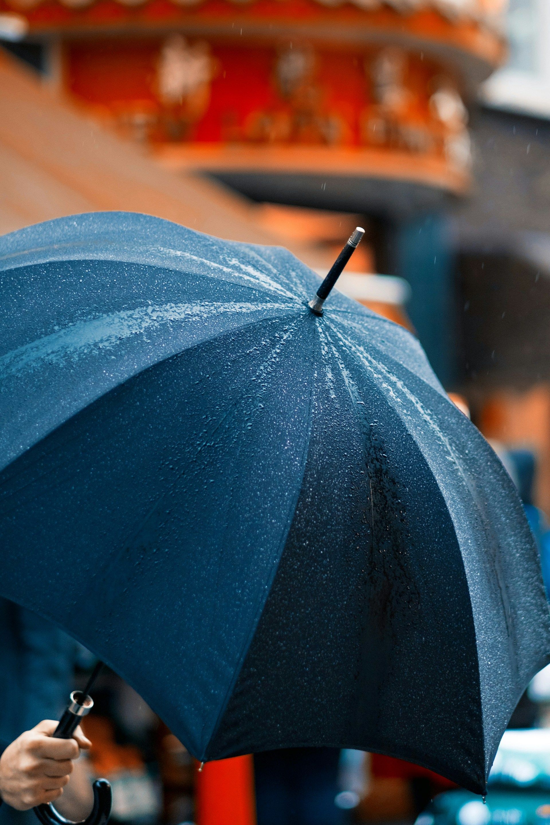A dark blue umbrella covered in raindrops held by a hand, with a blurred outdoor scene in the background.