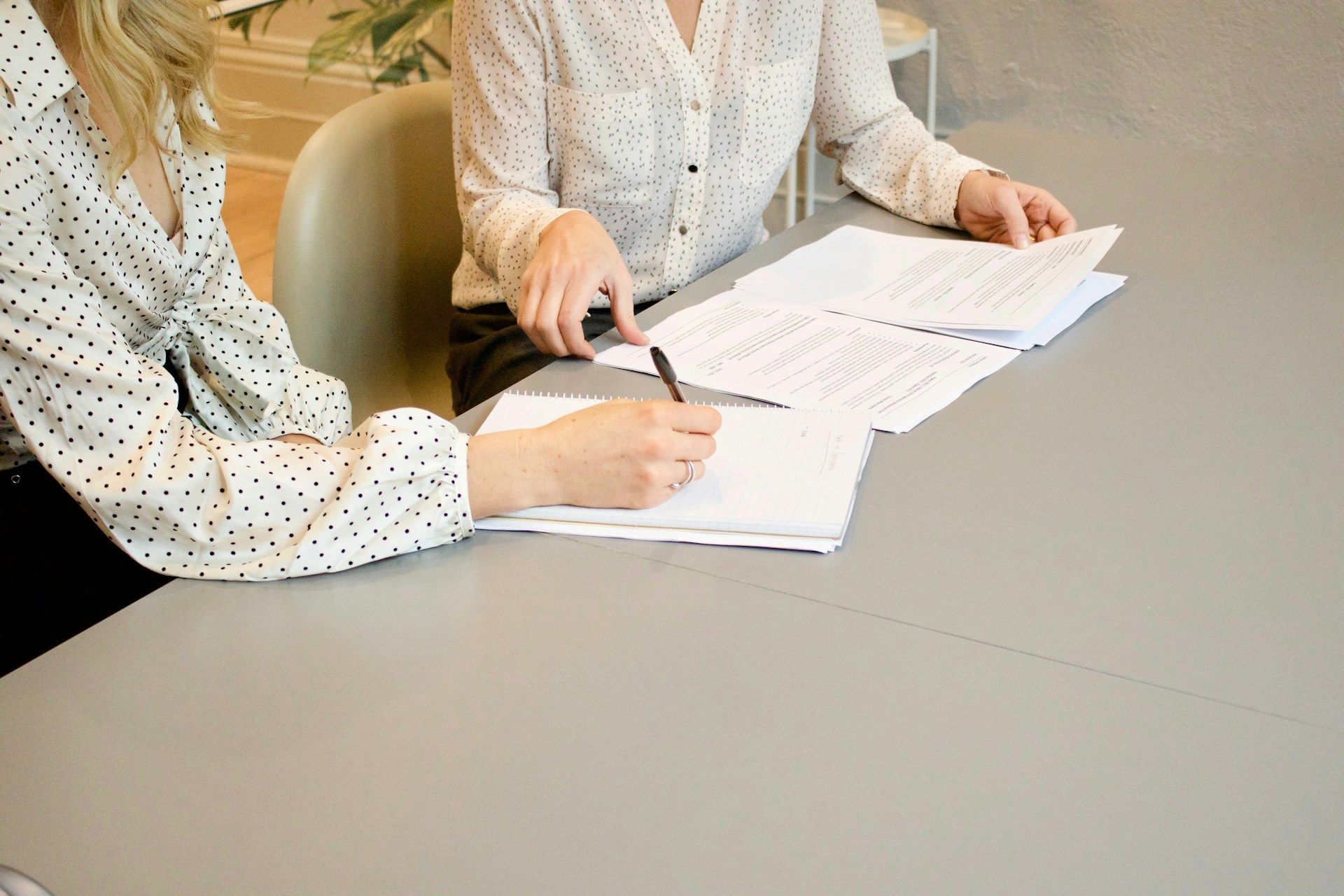 Two people in white polka-dot blouses sit at a table, reviewing and writing on documents together in an office setting.