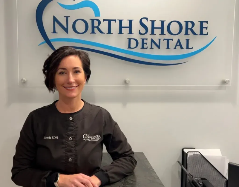 Woman in dark uniform seated in front of North Shore Dental sign, smiling in an office.