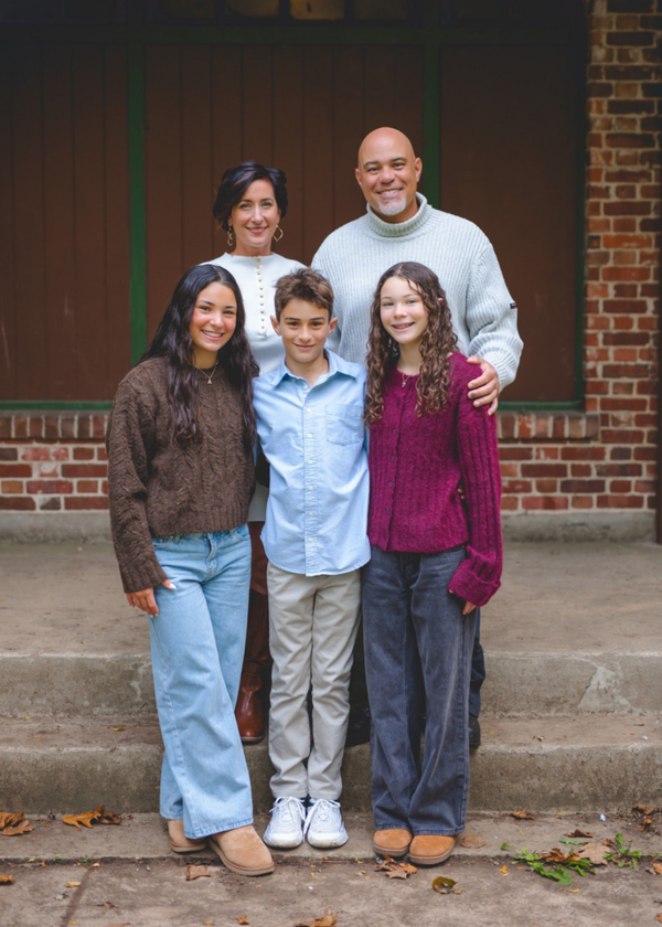 Family of four standing on front steps in front of a brick house, smiling at the camera.