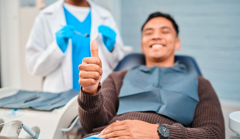Smiling dental patient giving thumbs-up while reclining in a chair, with dentist standing behind him.