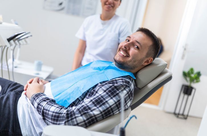 Smiling patient reclines in a dental chair while a dentist stands nearby in a bright clinic.