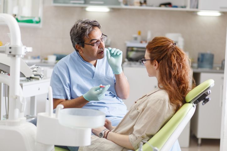 Dentist consulting with a seated patient in a bright dental clinic, holding a dental tool.