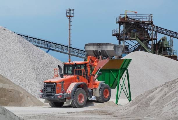 Front loader moving gravel at quarry site with heavy machinery and large aggregate piles working now.