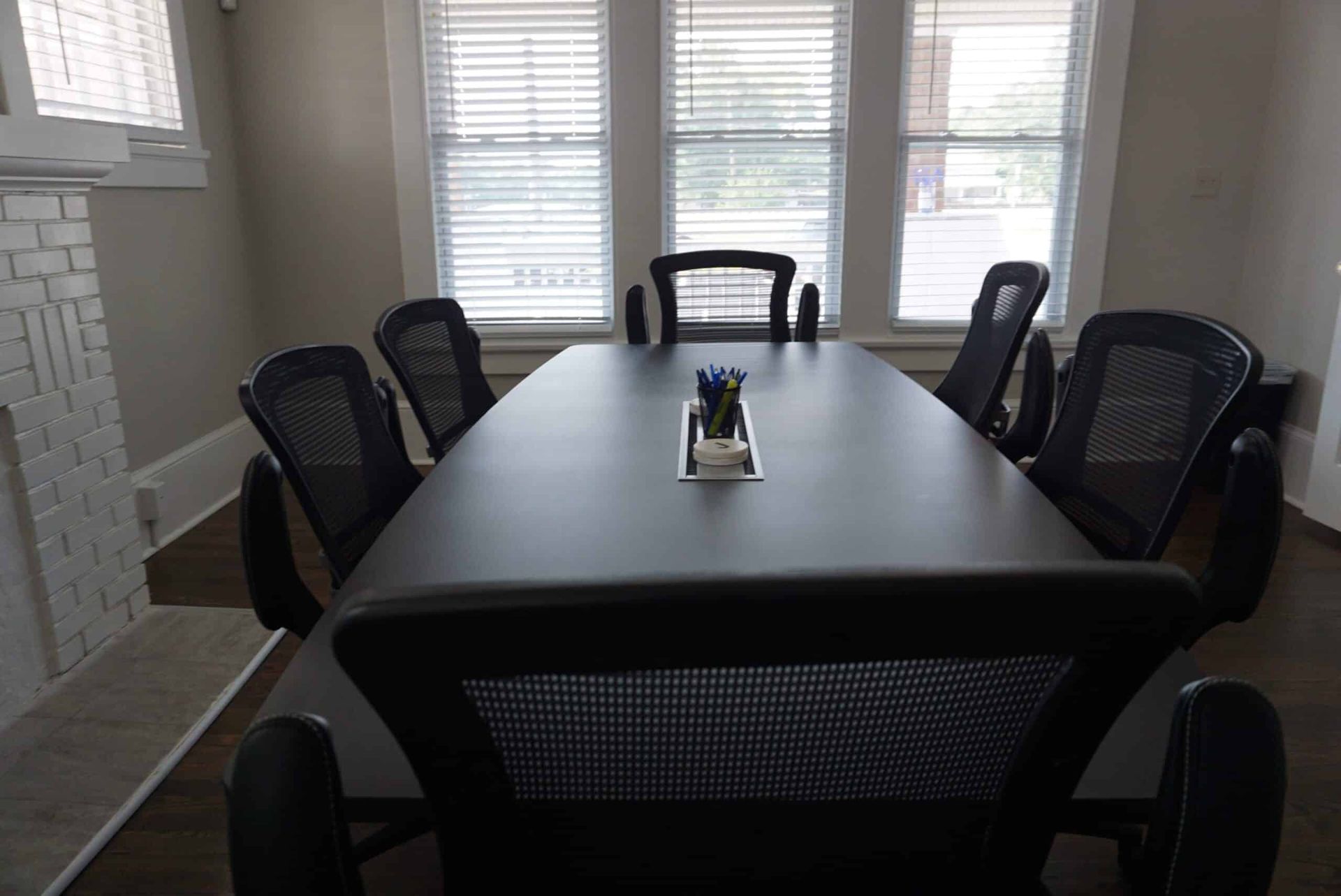 Conference room with a long black table, six black chairs, and bright windows in back.