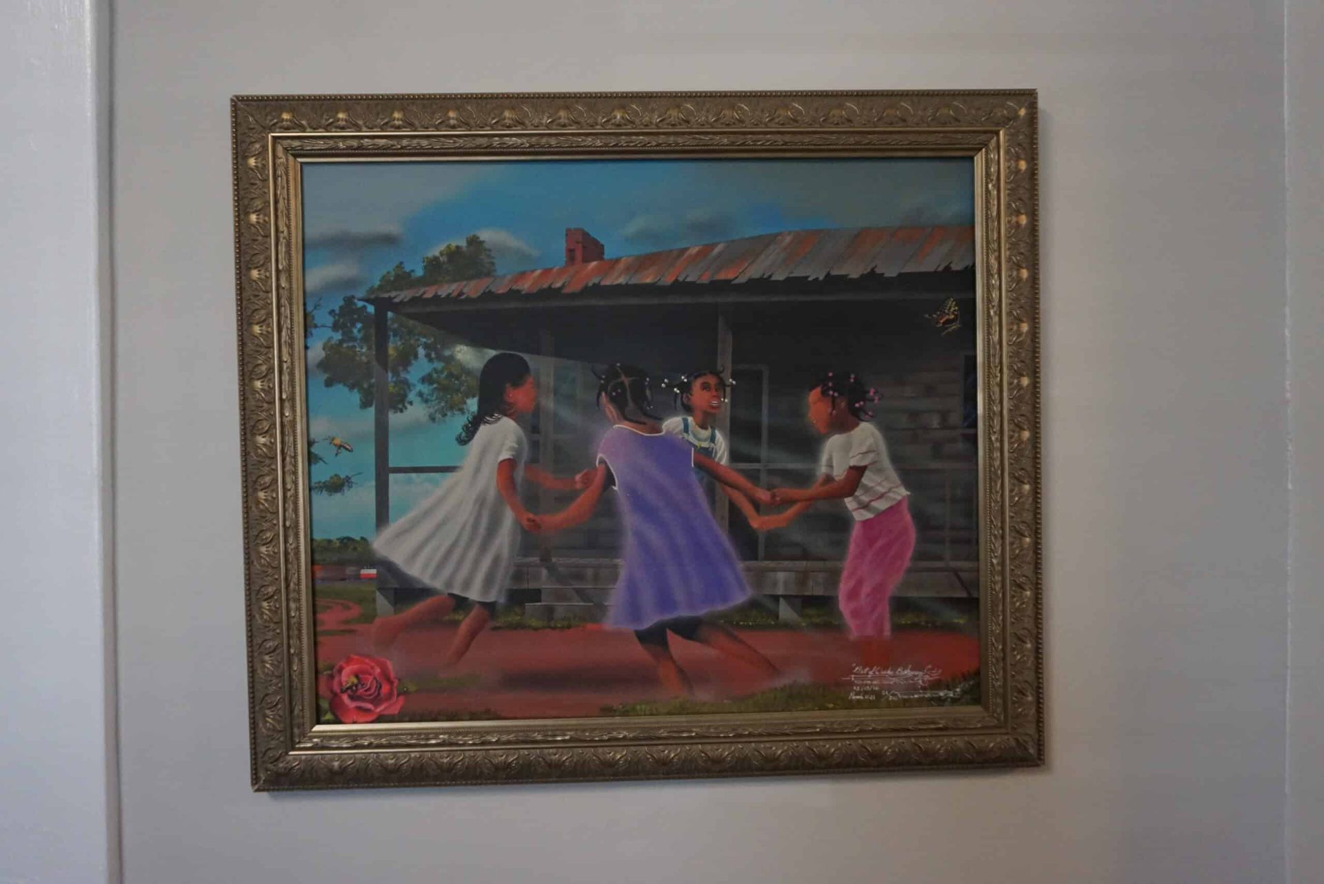 Framed painting of three people dancing outdoors in colorful dresses, with a house in the background