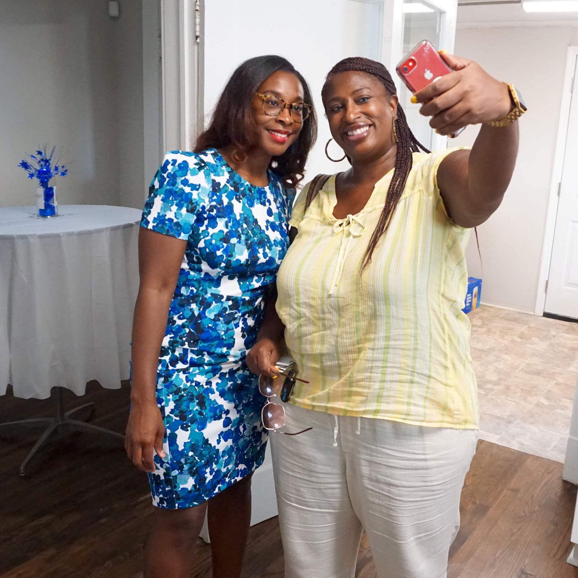 Two women smiling and posing for a selfie indoors, one in a yellow top and one in a blue floral dress.
