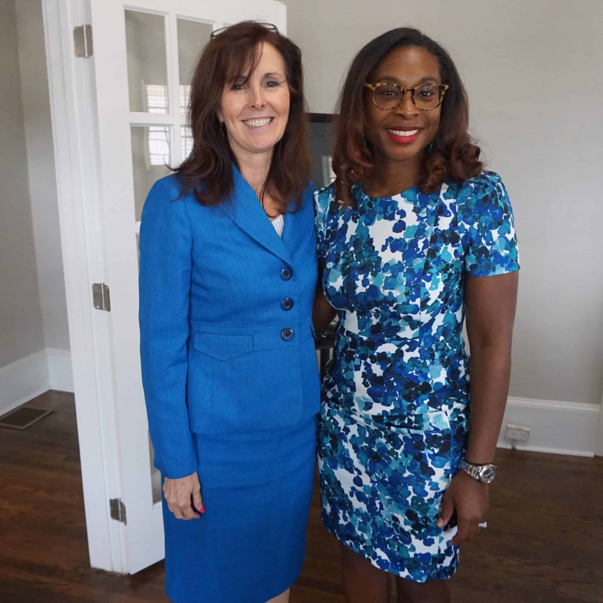 Two women smiling indoors, wearing blue dresses, standing together near a doorway