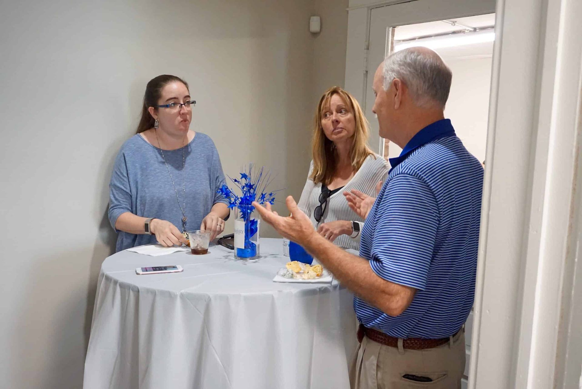 Three people chatting around a small round table in a bright hallway or office.
