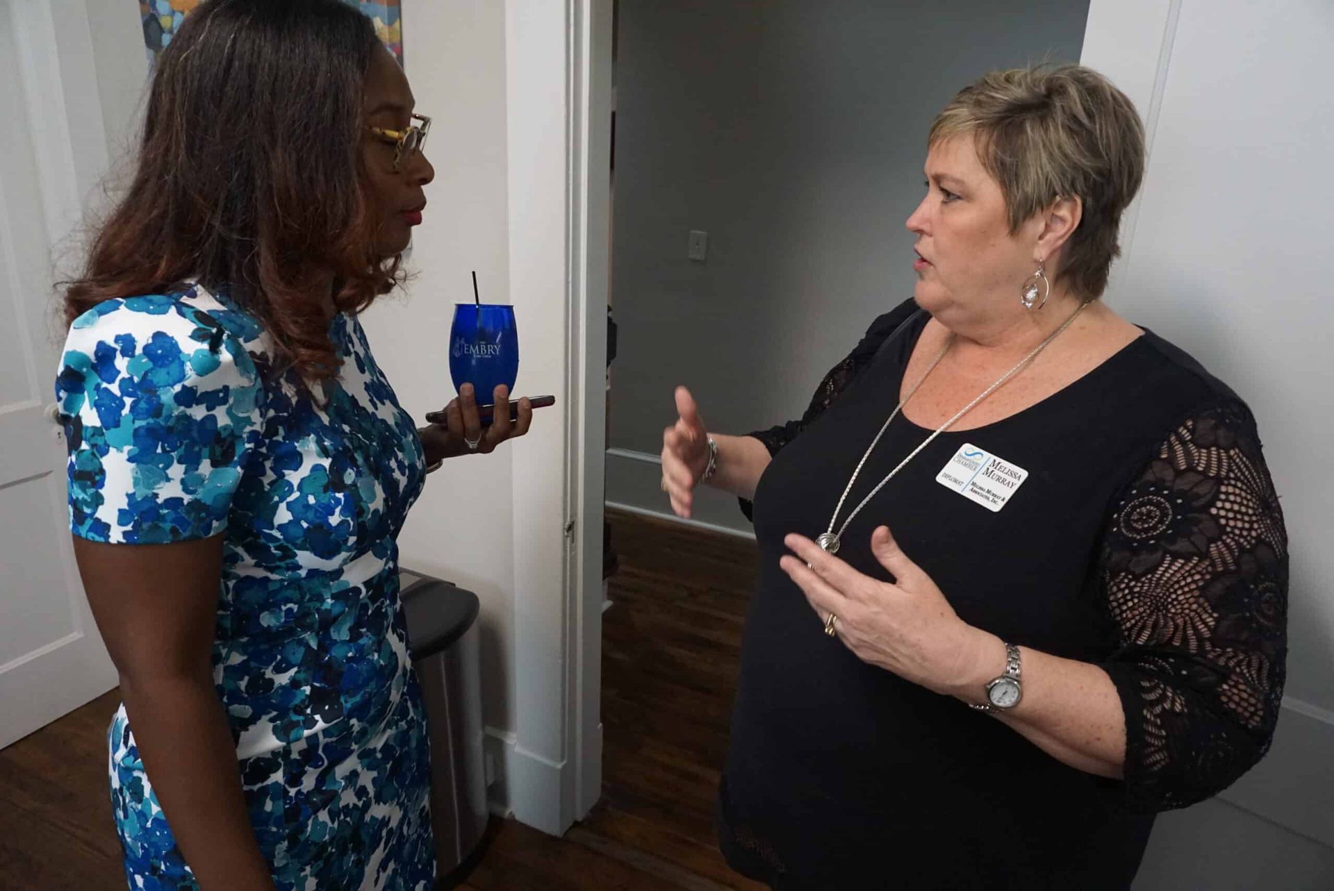 Two women talking in a doorway, one holding a blue cup and the other gesturing with both hands.