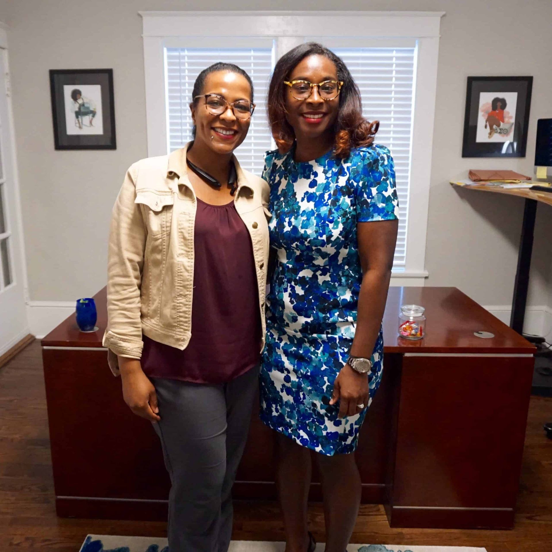 Two people smiling and posing in an office with a wooden desk and framed wall art.