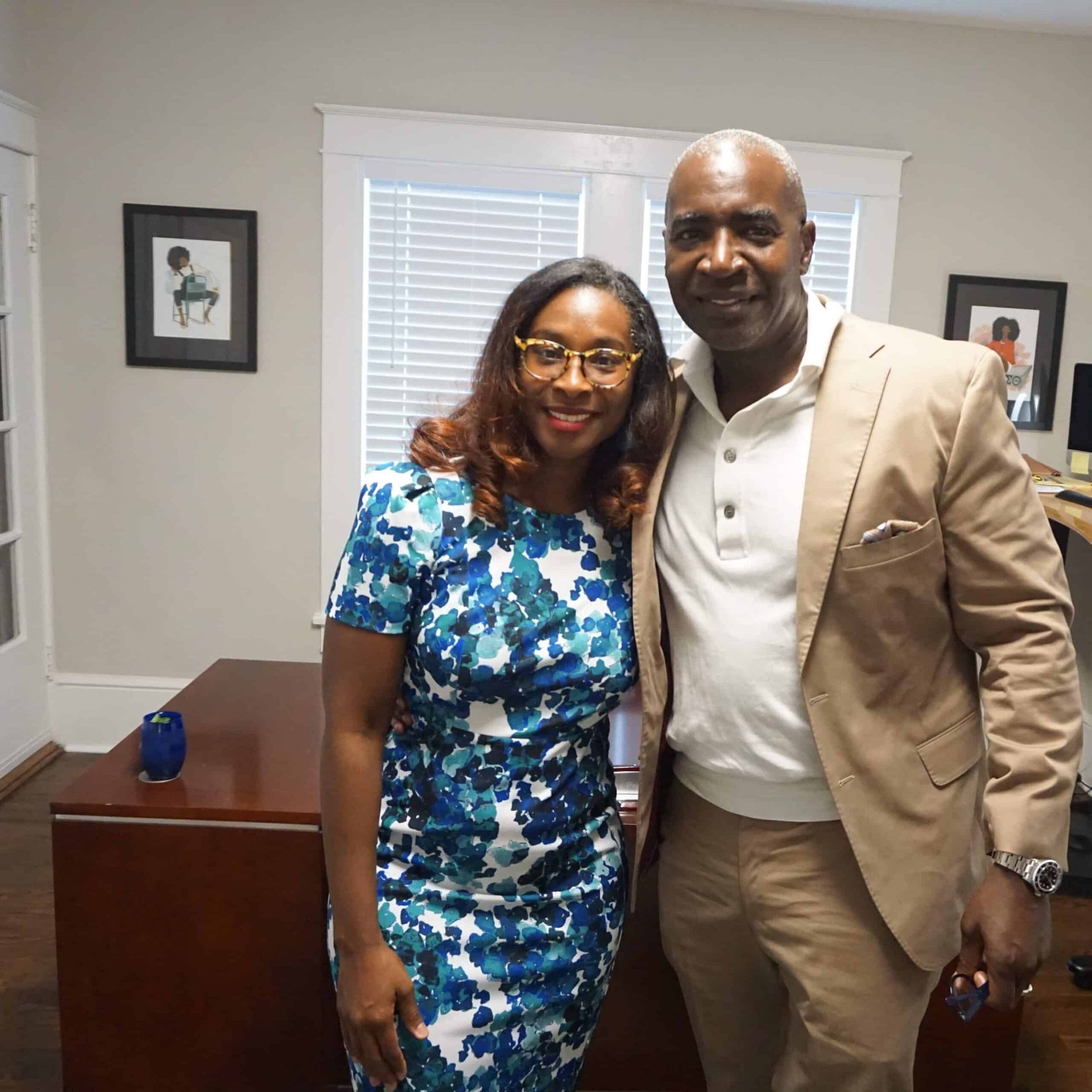 Two people smiling indoors, standing beside a desk in an office.