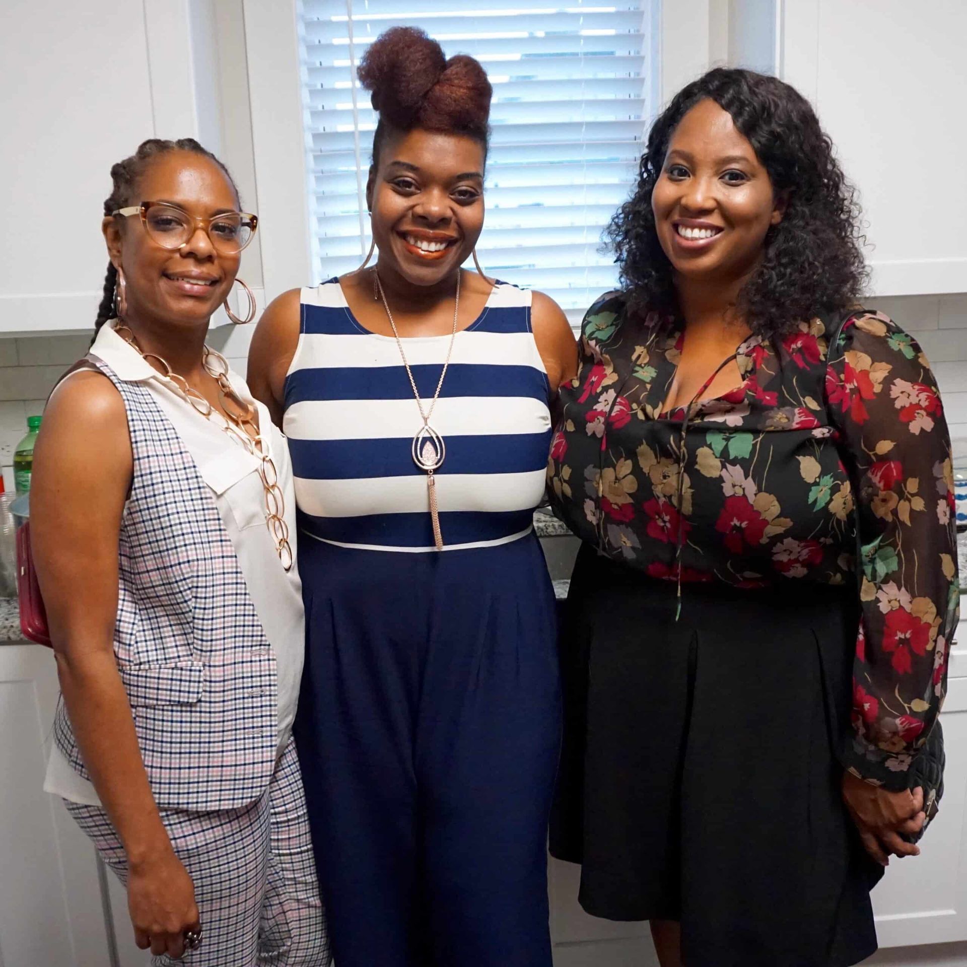 Three women smiling indoors, standing close together in front of a window.