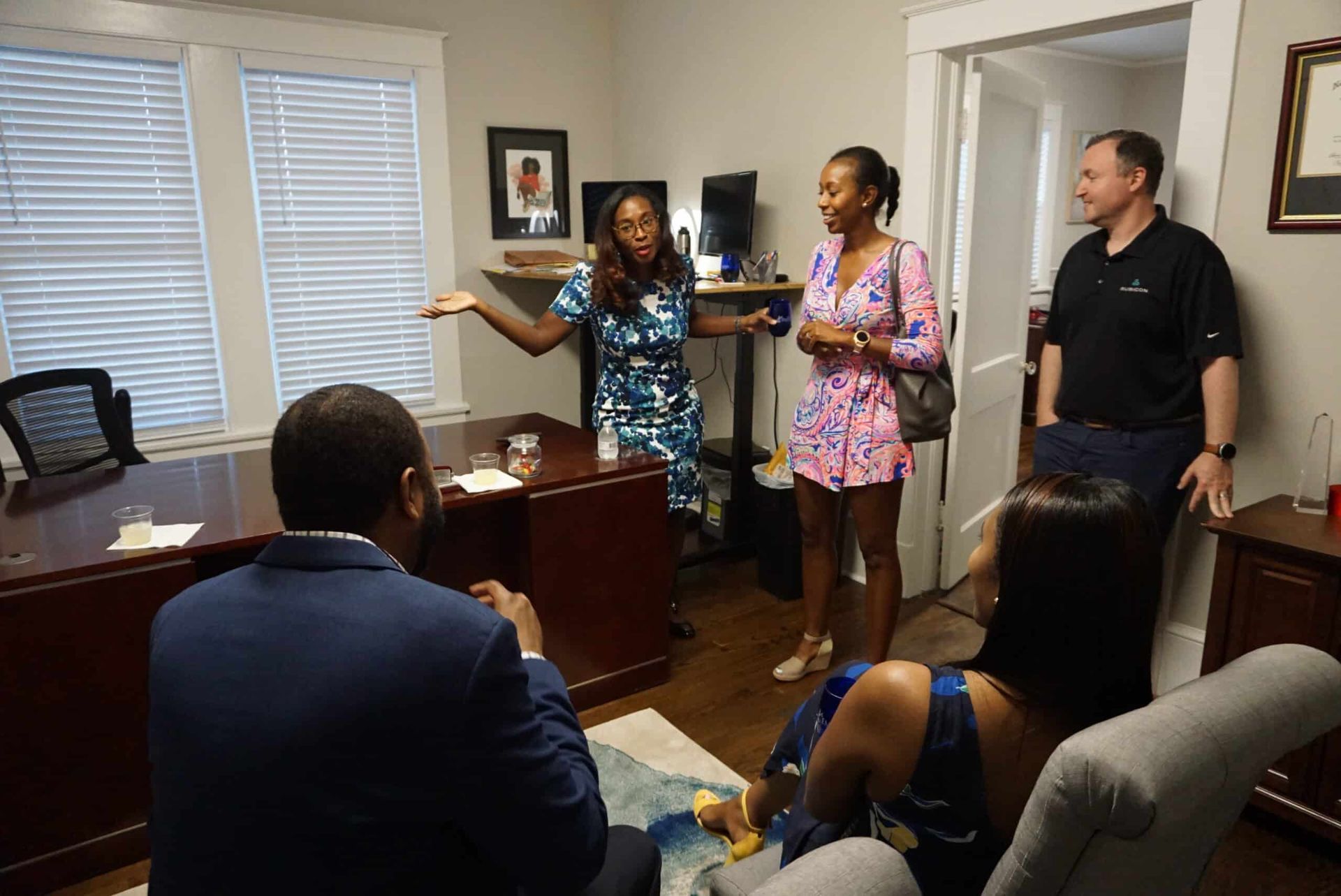 People in an office meeting, one woman speaking while others listen around a desk.