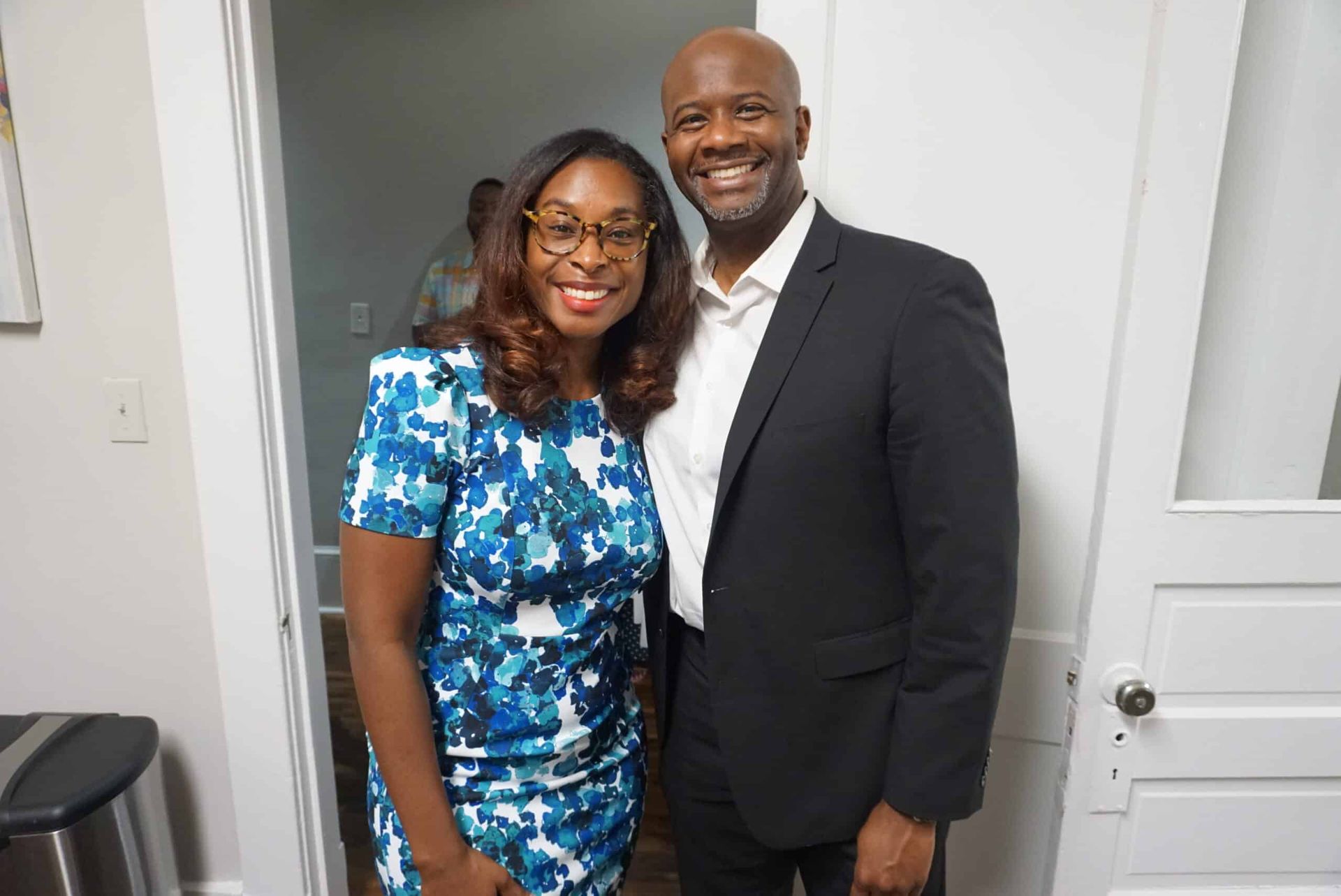 Two people smiling indoors, standing together in a doorway, one in a blue floral dress and one in a dark suit.
