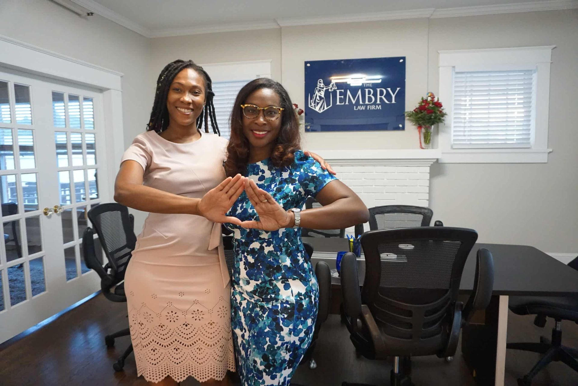 Two women posing in an office, smiling and making a heart shape with their hands.