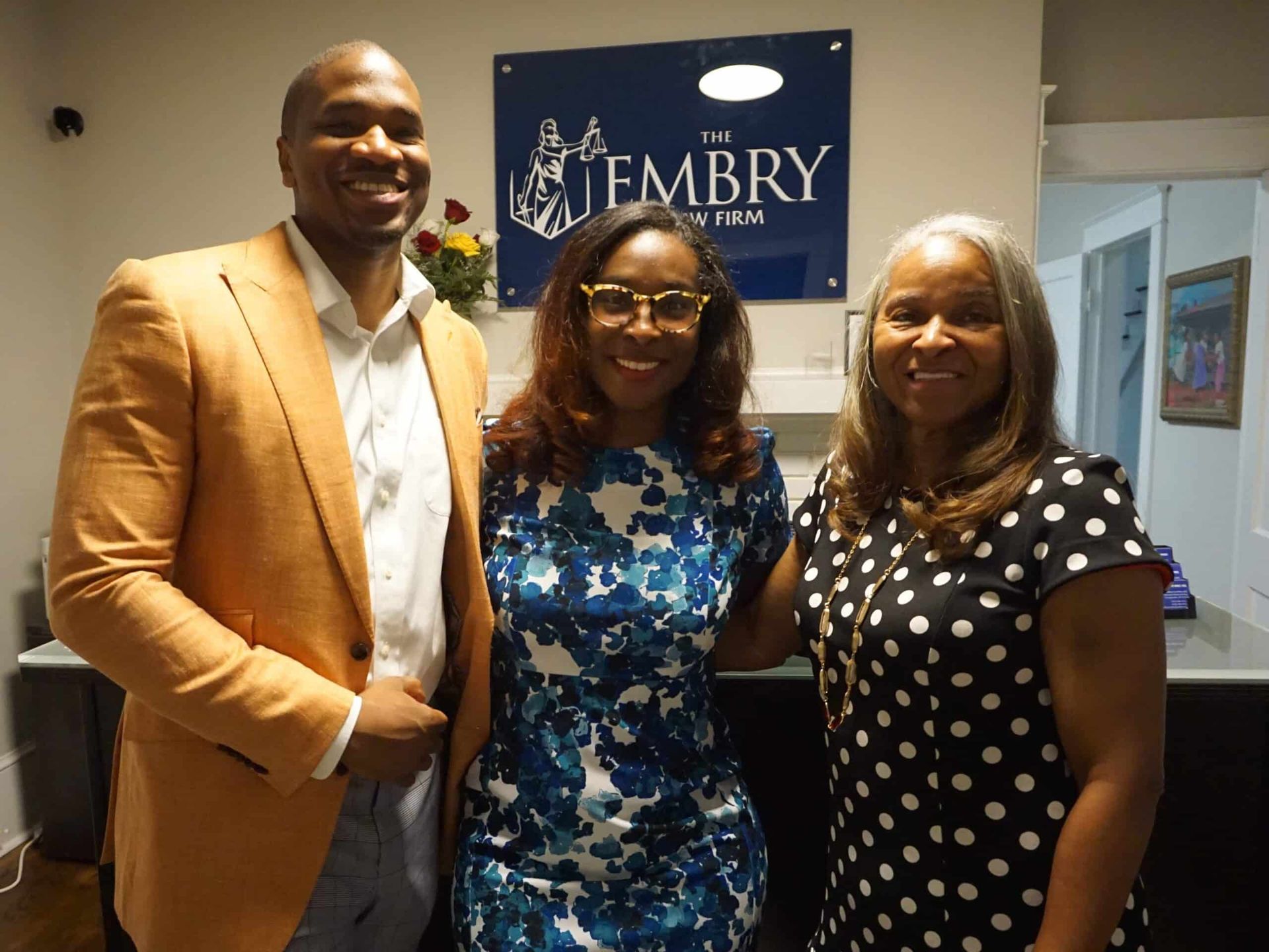 Three people smiling and posing indoors in front of a Jembry sign.