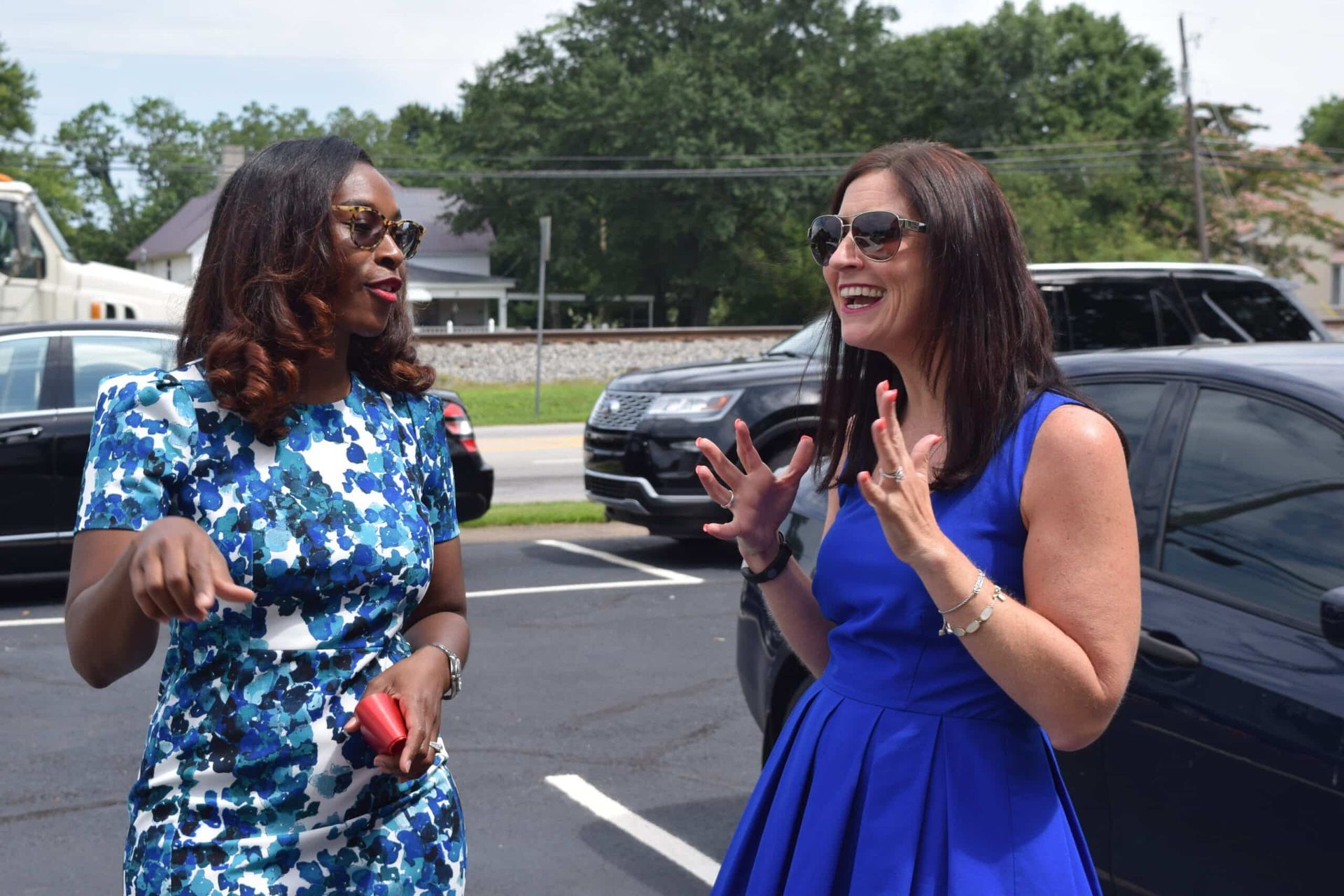 Two women talking and smiling in a parking lot, one in a blue dress and one in a blue floral outfit.