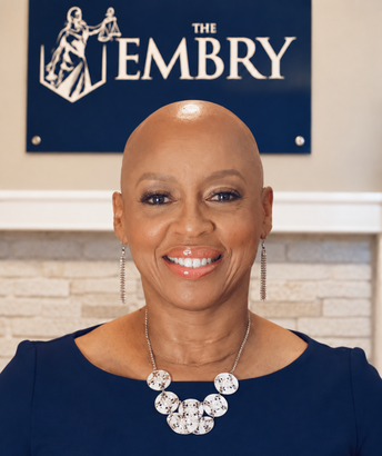 Smiling person in navy top stands before The Embry sign in a bright interior.