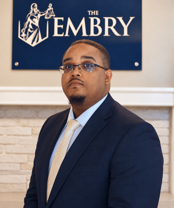 Man in a navy suit and tie standing indoors beneath a sign reading “The Embry.”