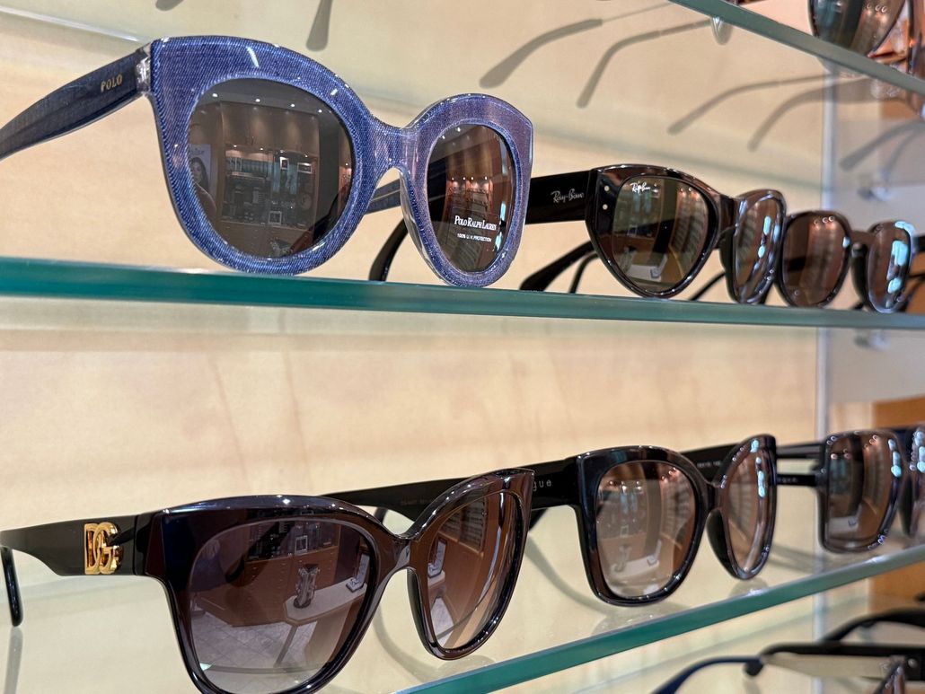 Occhiali da sole esposti su scaffali di vetro, tra cui modelli con montatura blu e marrone, in un negozio.