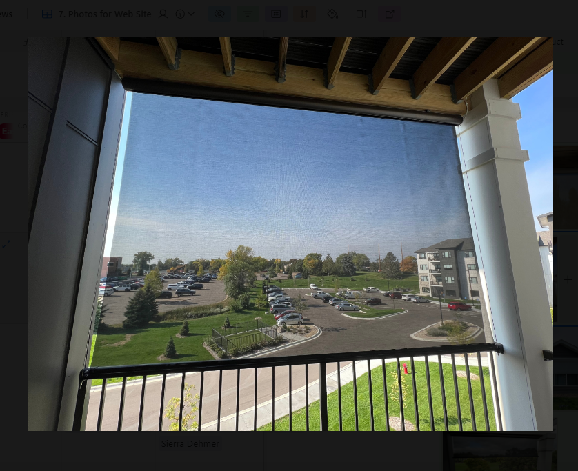 Balcony with retractable sun shade overlooking parking lot, green space, and blue sky.