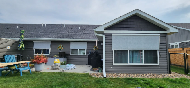 Backyard patio with a table, chairs, and grill under a cloudy sky. The house has gray siding and window shades.