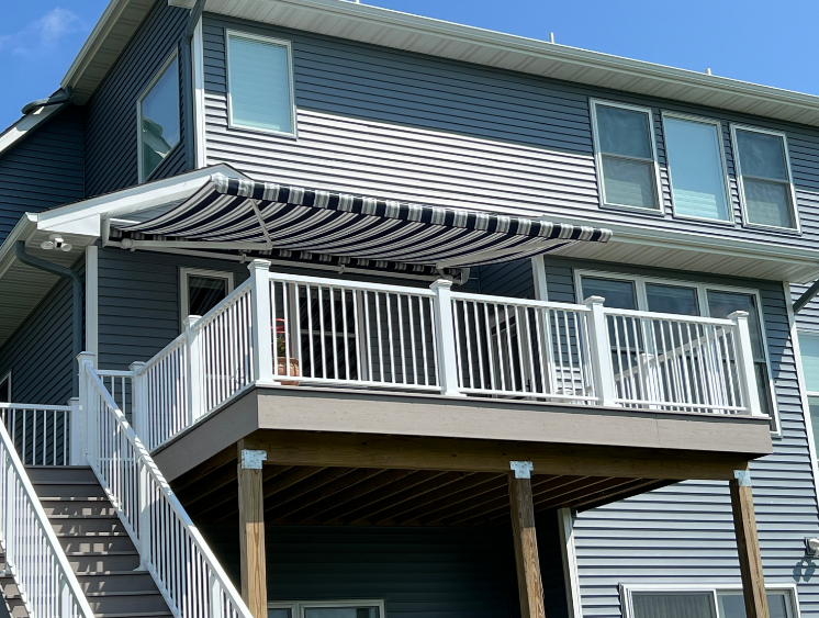 Two-story house with blue siding, white deck, and a black and white striped awning.