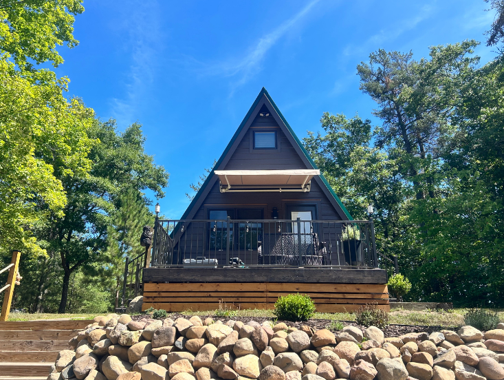A-frame cabin with brown exterior, wooden deck, and awning, surrounded by trees under a blue sky.