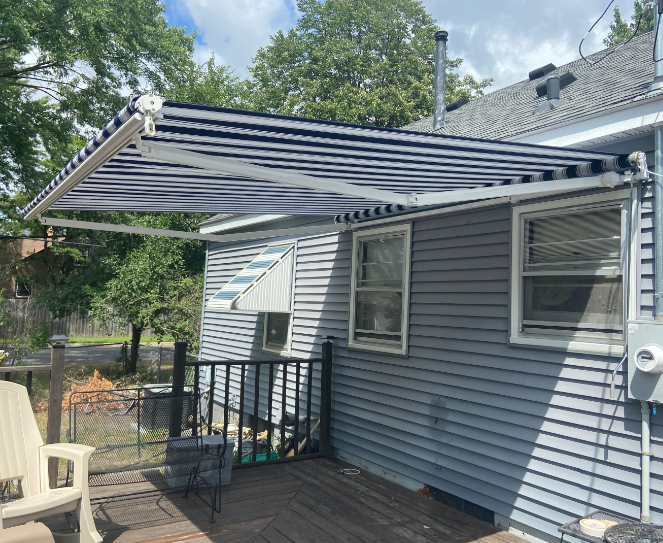 A retractable awning with blue and white stripes extends over a wooden deck attached to a gray house.