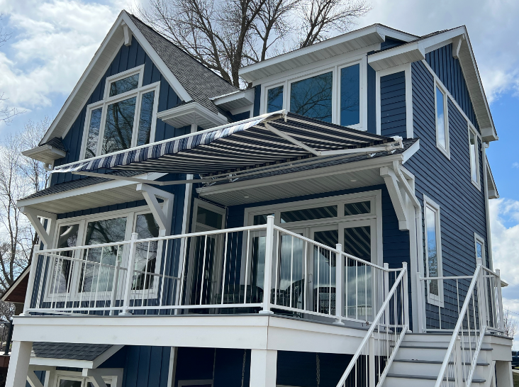 Three-story blue house with white trim, glass railing balconies, and a striped awning; blue sky.