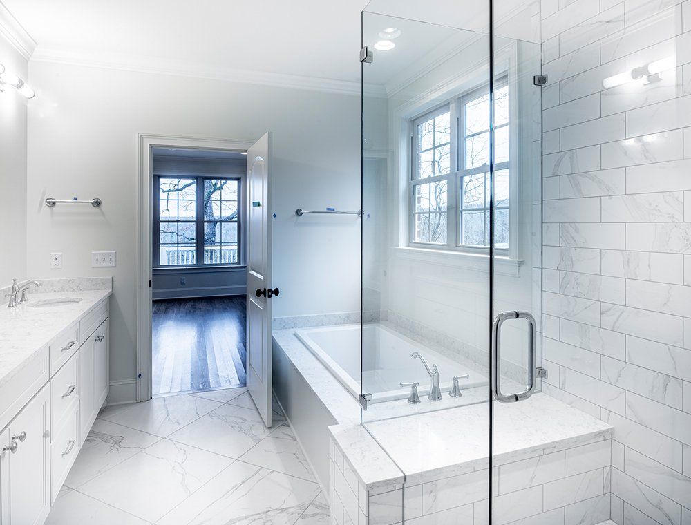 Bright, white bathroom with marble tile and glass shower. Bathtub, window, and open doorway to another room are also visible.