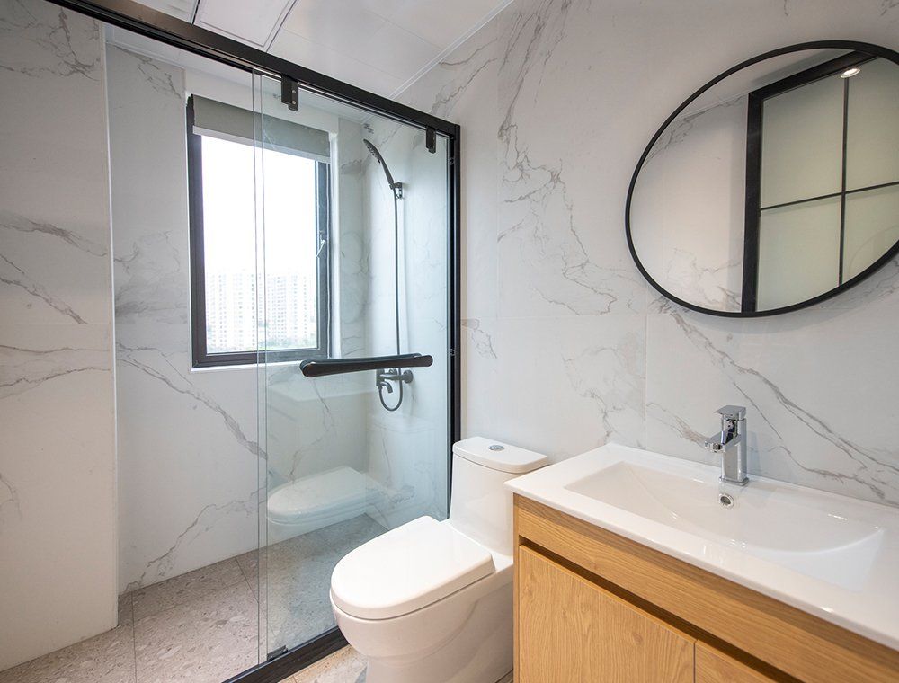 Modern bathroom with marble walls, glass shower, toilet, wooden vanity, and round mirror.