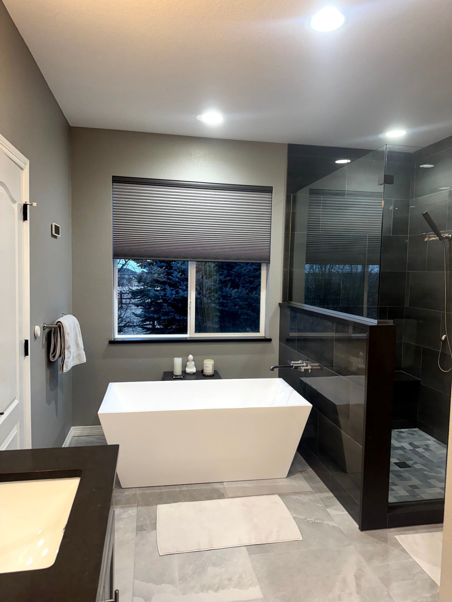 Modern bathroom with a white bathtub, dark shower, and patterned window shade.