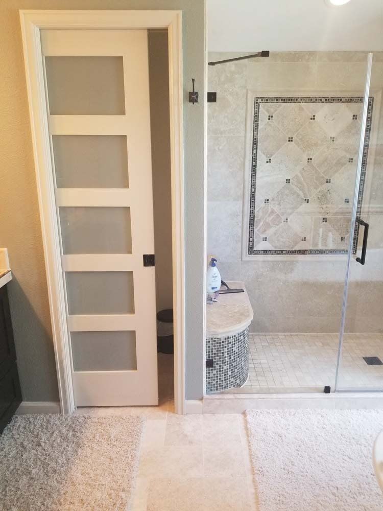 White door with frosted glass panels leads to a bathroom. Shower with glass door, bench, and patterned tile.