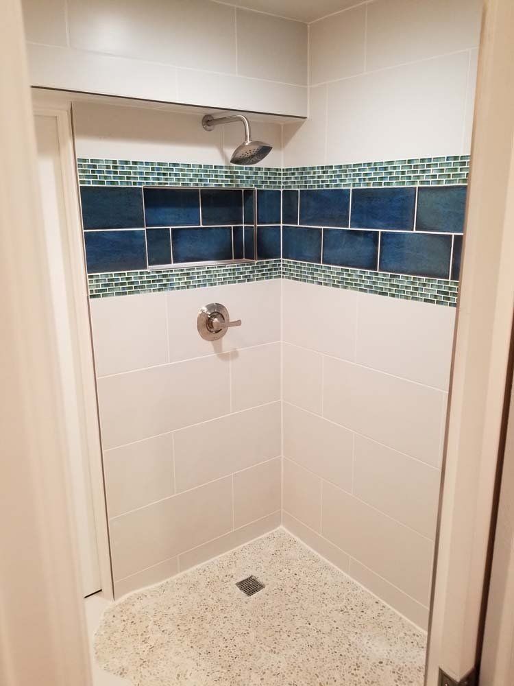 Small shower with white and blue tiles, a built-in shelf, and a speckled floor.