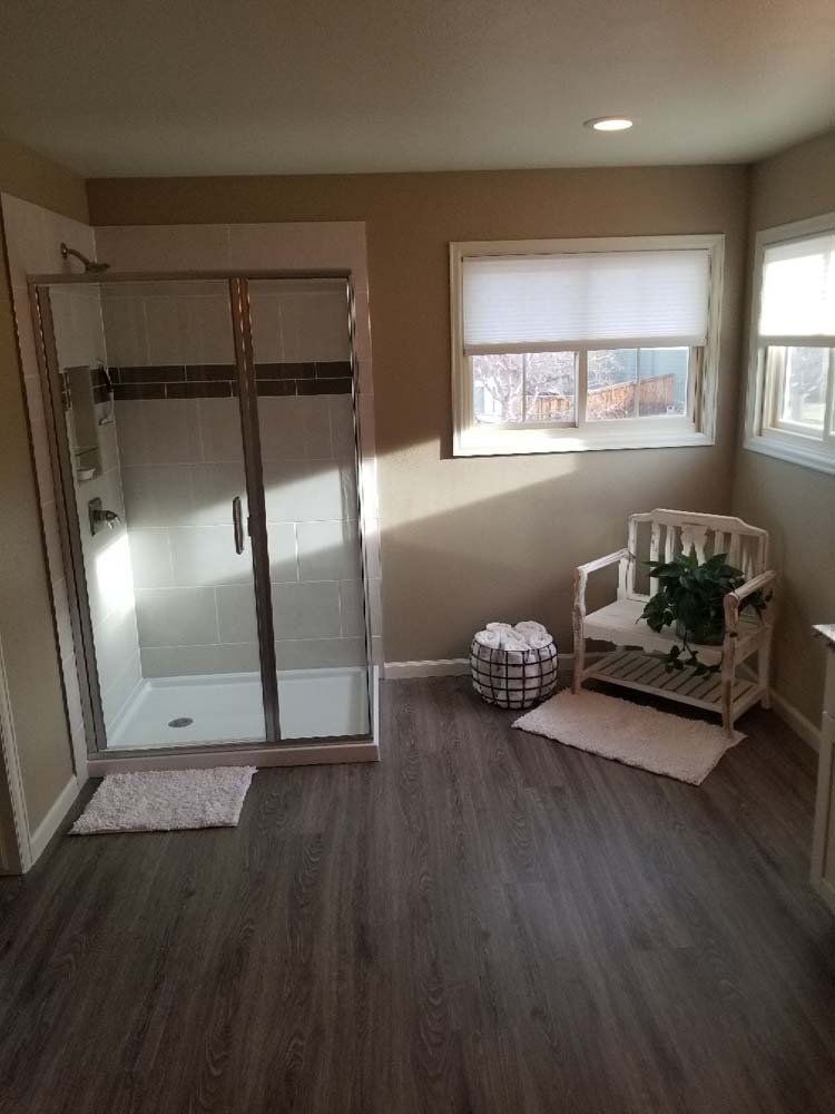 Bathroom with a glass shower, two windows, and a small chair. Gray floor and neutral walls.