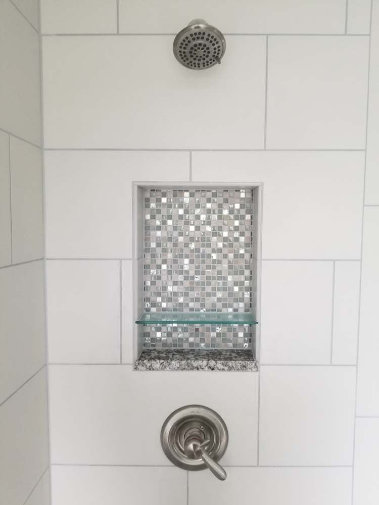 White tiled shower with built-in shelf, mosaic tile, glass shelf, and shower fixtures.