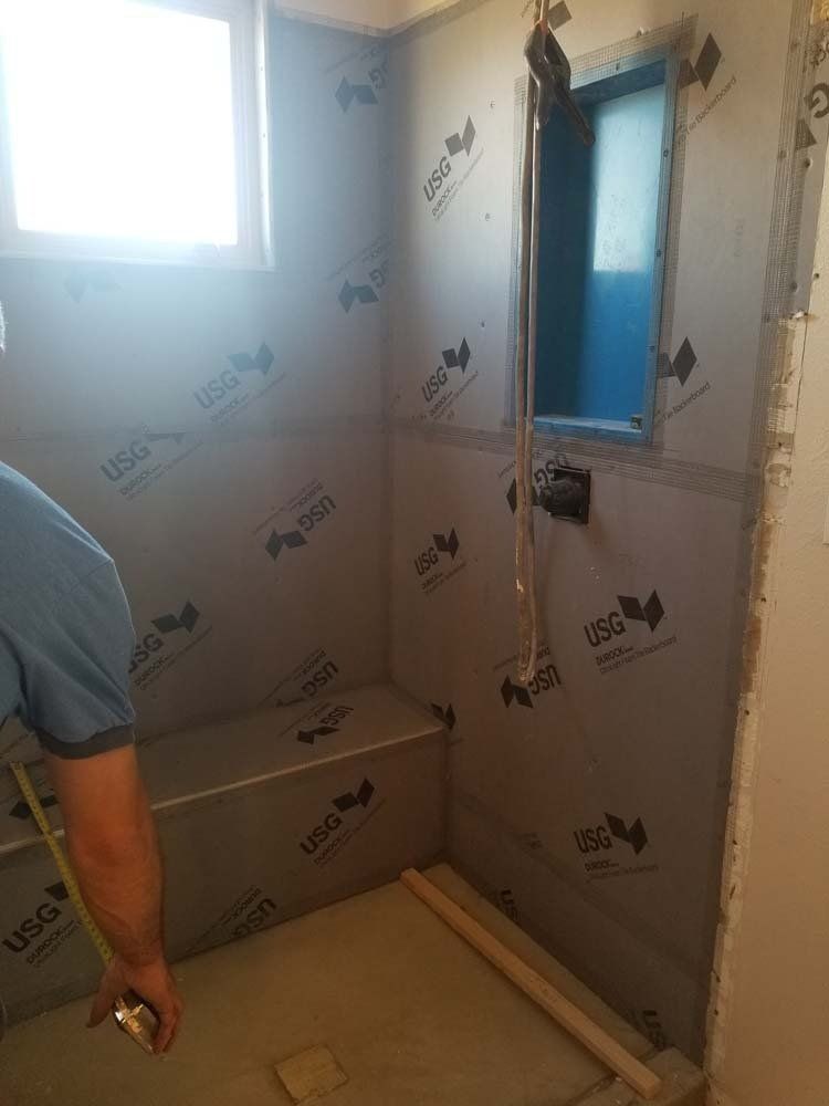 Bathroom under construction. Man measuring near a built-in bench. Gray waterproof panels cover the walls. Small blue window.